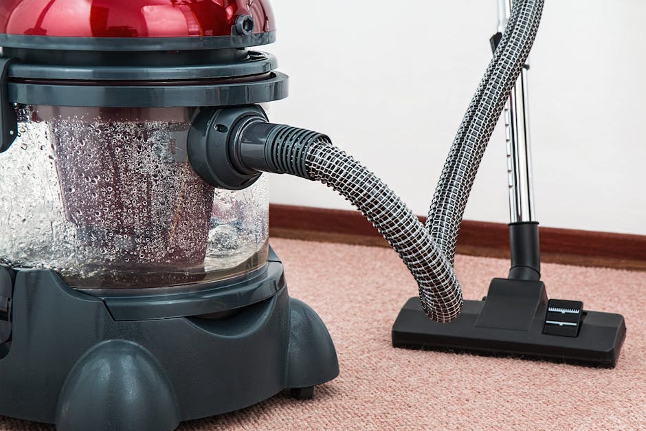 Close-up view of a person vacuuming a light oak hardwood floor in a residential room, with a canister vacuum cleaner connected to a flexible hose. The individual is wearing dark shorts, a long-sleeve 