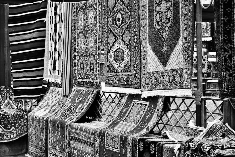 The image shows a collection of colorful, intricately patterned rugs hanging on a dark wooden outdoor wall, with some draped over a horizontal wooden bar. The rugs feature various traditional geometri
